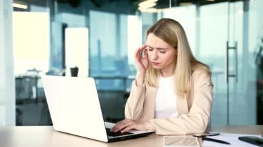 Modern ofiste çalışırken dizüstü bilgisayarla çalışırken baş ağrısı çeken yorgun genç bir iş kadını. Üzgün sarışın kadın acı hissediyor, masaj yapıyor ve elleriyle şakaklarını ovuyor.