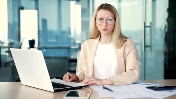一个年轻的 面带微笑的女商人坐在一个现代化办公室的办公桌前的画像 戴眼镜快乐的金发女员工工作 在电脑上打字 看着相机 — 图库视频影像