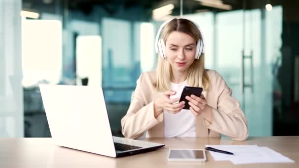 快乐的金发女人坐在现代办公室的工作场所 戴着耳机听音乐 一个笑容满面的女员工在智能手机上打开播放列表 欣赏音乐 — 图库视频影像