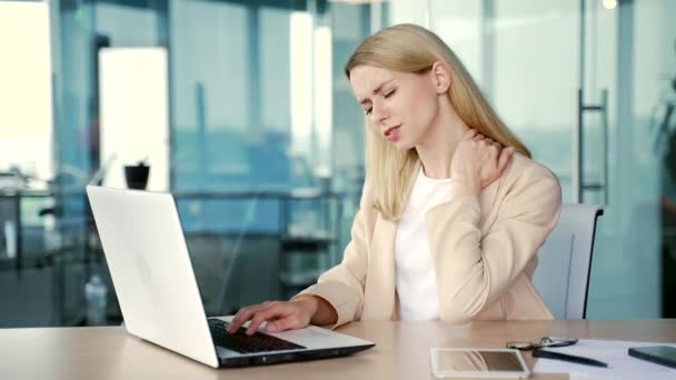 疲れたブロンドの実業家は 現代のオフィスの職場でデスクに座っている間 ノートパソコンで作業しながら首の痛みに苦しんでいます スーツのマッサージで悲しい女性の従業員と筋肉をこすり ストレッチ — ストック動画