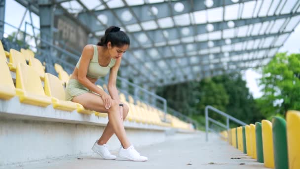 若いスポーティな女性は 都市のスタジアムに座っている間 痛みを伴うひざ関節痛に苦しんでいます 女性のマッサージストレッチ ジョギング中に外傷 筋肉クランプ スプレイン カーフ スポーツ — ストック動画