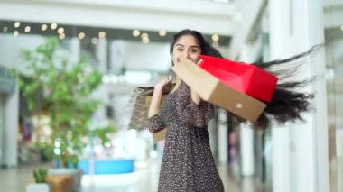 Satış ve indirimlerden sonra alışveriş merkezinde alışveriş yapan mutlu genç Hintli kadın renkli hediye çantalarıyla gülümseyen ve içeriden satın alan memnun alıcıyla kameraya bakıyor.