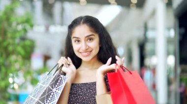 Satış ve indirimlerden sonra alışveriş merkezinde alışveriş yapan mutlu genç Hintli kadın renkli hediye çantalarıyla gülümseyen ve içeriden satın alan memnun alıcıyla kameraya bakıyor.