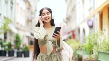 Neşeli Hintli genç Arap kadın, şehir sokaklarında yürüyüp mutluluk ve yaşam tarzı kavramı ile dans eden neşeli kız kıza yürürken akıllı bir telefonla müzik dinliyor.