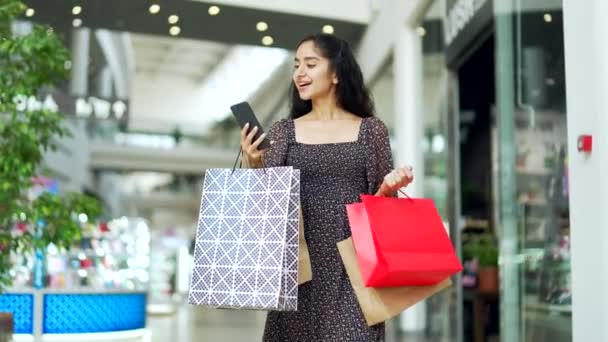 若い美しい幸せな女性の買い物客の喜びの顧客はショッピング センター モールを歩き スマートフォンを入力する携帯電話のブラウジング チャット ギフトの色のパッケージ袋と満足して店に行く — ストック動画