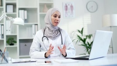 Hastane kliniğinde laptopla konuşan kendine güvenen Müslüman kadın doktor. Kadın tıp doktoru, ofisteki çalışma masasında oturan bir hastayla sohbet yardımı alıyor.