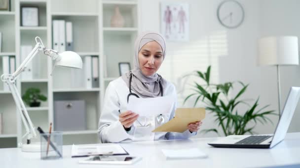 ハッピーイスラム教徒の女性医師は 現代の病院の診療所で職場に座っている素晴らしいニュースを受け取りました 楽しい女性の医療従事者医師は快適な通知で喜びます — ストック動画