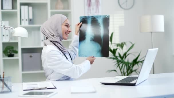 穆斯林女医生一边拿着一台笔记本电脑一边用视频通话 一边在一家现代化的医院诊所里进行X光检查 医务工作者女医生咨询在职工作场所的病人 — 图库视频影像