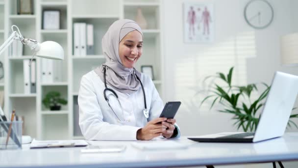 快乐的穆斯林女医生使用浏览智能手机 在现代医院诊所输入讯息 微笑的女医务工作者在办公室的办公桌前给病人发短信 — 图库视频影像