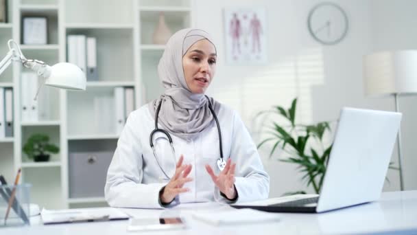 有自信的穆斯林女医生在医院诊所用手提电脑进行视频通话 女医务工作者医生正在与办公室里坐在办公桌前的一位病人进行谈话 — 图库视频影像