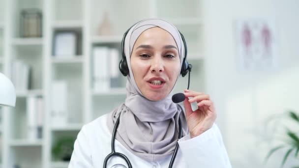 摄像头的视图 在医院诊所用无线耳机对着视频通话的女医生微笑 快乐医疗工作者的医生会诊 帮助病人在办公室里看着相机 — 图库视频影像