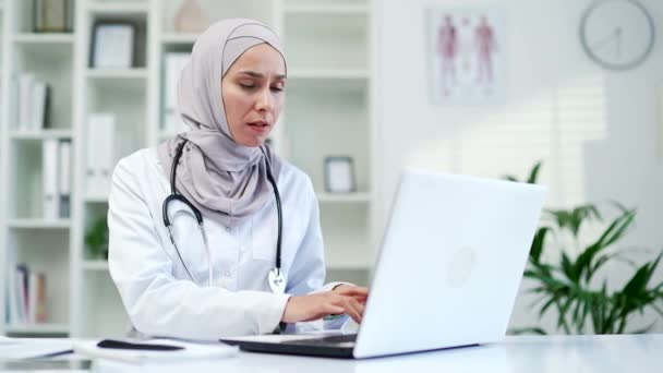 自信的穆斯林严重的女医生在笔记本电脑上浏览 并思考如何解决一个问题 而坐在办公桌医院诊所 在办公室输入计算机的女医生 — 图库视频影像