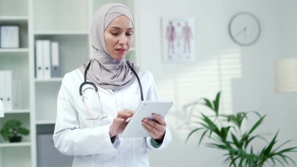 一名身穿白衣的穆斯林女医生在一家现代医院诊所的平板电脑上使用浏览打字 医生在办公室工作时正在和一位病人交谈 — 图库视频影像