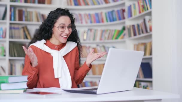 若い幸せなメガネの女性教師は キャンパス図書館のスペースでラップトップコンピュータを使用してオンラインビデオ通話トレーニングを持っています 大学や大学のチューターがウェビナーを始める レッスン 教室のミーティングがあります — ストック動画