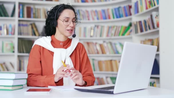 ヘッドセットの若い幸せな女性教師は キャンパス図書館のスペースでラップトップコンピュータを使用してオンラインビデオ通話トレーニングを持っています 大学や大学のチューターがウェビナーを始める レッスン 教室のミーティングがあります — ストック動画