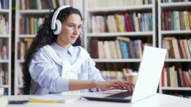 Kulaklıklı kız öğrenci, kampüs kütüphanesinde öğrenmek için dizüstü bilgisayar kullanıyor. Kız bilgisayarda çalışıyor, üniversitenin sınav dönemi için hazırlanırken internette geziniyor.