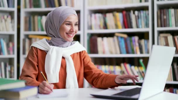 穆斯林女学生坐在学校图书馆时 在笔记本电脑上做笔记 女生在考试期间用电脑浏览 检查作业 准备考试 — 图库视频影像