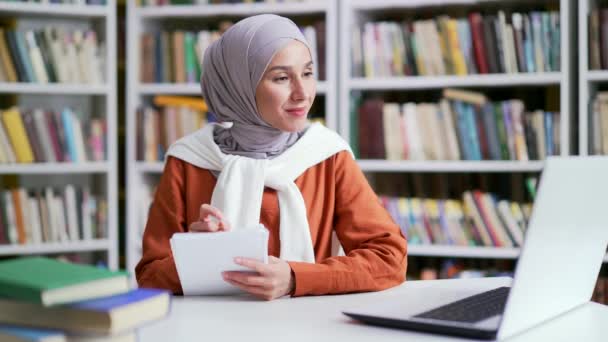 带着笔记本 头戴头巾的穆斯林女学生在校园图书馆使用笔记本电脑 通过视频电话收听在线电子学习 在大学的网络研讨会上观看远程学习研讨会 — 图库视频影像