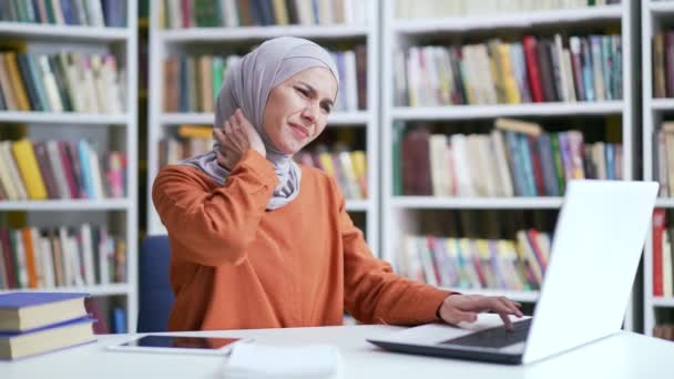 ヒジャーブのムスリム女性学生は キャンパス図書館のスペースに座っている間 ノートパソコンで作業しながら首の痛みに苦しんでいます 疲れた女の子はマッサージし 筋肉を痛めます 彼女は過剰になっているか 冷たい神経を持っています — ストック動画
