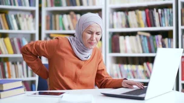 穆斯林女学生在校园图书馆的笔记本电脑上工作时背痛 女孩按摩下背部 按摩脊椎肌肉 她有坐骨神经痛或冷神经 — 图库视频影像