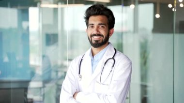 Modern hastane kliniğindeki kameraya bakan beyaz önlüklü, gülümseyen sakallı bir tıp doktorunun portresi. Ofiste kollarını kavuşturmuş pozitif tıp doktoru pozu