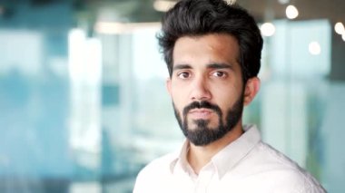 Kapatın. Modern bir ofiste duran beyaz gömlekli, sakallı, genç bir girişimcinin portresi. Güvenilir başarılı iş adamı kameraya bakarak poz verir. Müdürün ya da çalışanın vesikalık fotoğrafı.