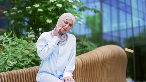 ヒジャーブの若いイスラム教徒の女性従業員は オフィスビルの近くの通りのベンチに座っている間に歯痛に苦しんでいます 痛みの場所をマッサージする女性 彼女は歯医者の助けを必要としています — ストック動画