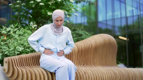 ヒジャーブの病気の若いムスリム女性従業員は オフィスビルの近くのベンチに座っている胃の痛みに苦しんでいます 女性は胃炎 消化器系の問題 便秘または肝臓を持っています — ストック動画