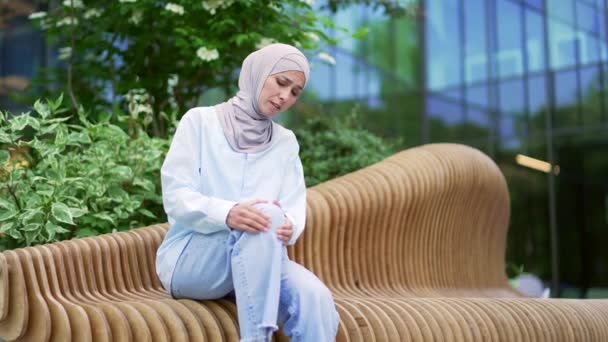 在写字楼附近的一条长椅上 年轻的穆斯林女员工坐在街上时 膝关节疼痛 疼痛严重 悲哀的女人按摩腿部肌肉 风湿病 — 图库视频影像