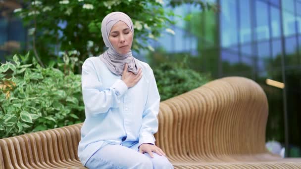 胸の張力に苦しんでいるヒジャブの病気の若いイスラム教徒の女性従業員 オフィスビルの近くの通りのベンチに座って心臓発作 女性は胸の痛みを感じ 緊急の医療を必要とします — ストック動画