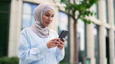 Mutlu, tesettürlü Müslüman kadın çalışan bir ofis binasının yakınındaki akıllı telefondan harika haberler okuyarak başarıyı kutluyor. Gülümseyen kadın neşeli bir mesaj aldı, evet işareti yaptı.