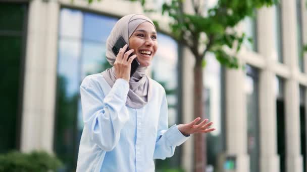 Młoda Uśmiechnięta Muzułmanka Hidżabu Rozmawia Przez Smartfona Stojącego Pobliżu Biurowca — Wideo stockowe