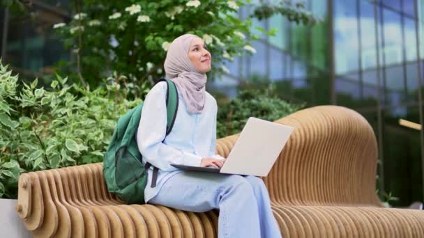ヒジャーブのムスリム女性学生は 大学ビルの近くのキャンパススペースのベンチに座っているラップトップにタイピングしています 遠隔学習セミナーの女の子 入学試験の準備をしています — ストック動画