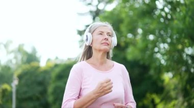Şehir parkında koşan yaşlı, gri saçlı kadın koşucu. Olgun, yaşlı kulaklıklı kadın sabahları müzik dinlemeyi ve doğada koşmayı sever. Yaşlı sporcu kadın spor yapıyor.