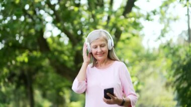 Kulaklıklı yaşlı, aktif, gri saçlı bir kadın şehir parkında müzik dinlemekten zevk alıyor. Mutlu, olgun, formda bir kadın. Çalma listesi ve antrenmandan sonra dinlenme de dahil.