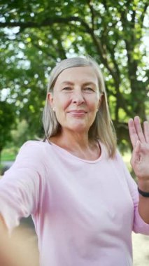 POV dikey video. Şehir parkında kameralı bir telefonla konuşan yaşlı, aktif, gri saçlı bir kadın. Yaşlı kadın kameraya bakıyor, arkadaşıyla antrenman hakkında olumlu izlenimler paylaşıyor.