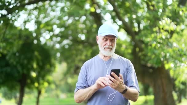 ヘッドフォンのシニアアクティブな灰色のひげ付き男は 都市公園を歩いている間にスマートフォンを使用します 成熟した古いフィットネス男性は オンラインでチャットしている ソーシャルネットワークをスクロールしたり 音楽を選択する — ストック動画