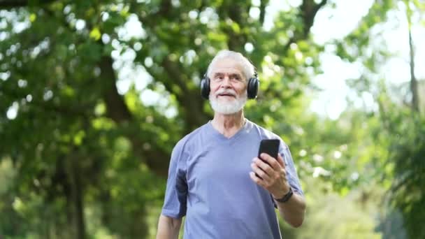 一个身披灰发 身披大胡子 头戴耳机的大个子男子在公园里一边走一边听音乐 健康的老运动员在训练或晨跑后放松快乐 — 图库视频影像