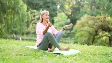 Yaşlı, aktif, gri saçlı kadın, şehir parkında bir paspasın üzerinde çıplak ayakla meditasyon yapıyor. Olgun, yaşlı kadın dışarıda yoga yapıyor. Emeklinin gözleri kapalı rahatlar