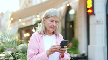 Yaşlı gri saçlı kadın şehir caddesinde akıllı telefon kullanıyor. Olgun yaşlı kadın cep telefonu çevrimiçi harita uygulaması, mesajlar yazmak veya okumak, çevrimiçi sohbetler veya sosyal ağlara göz atmak
