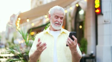 Yaşlı, gri saçlı, sakallı bir adam şehir caddesinde dikilirken akıllı telefon kullanıyor. Tişörtünü sallayan yaşlı ve mutlu bir erkek, telefon ekranına bakan bir arkadaşıyla konuşuyor.