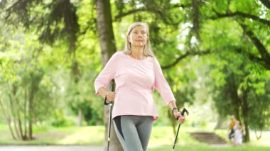 Şehir parkında yürüyüş direkleri olan yaşlı, gri saçlı bir kadın yürüyor. Olgun bir fitness kadını doğada İskandinav yürüyüşü yapıyor. Yaşlı sporcu kadın İskandinav yürüyüşü yapıyor.