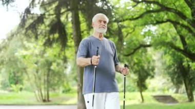 Şehir parkında yürüyüş direkleri olan yaşlı, gri saçlı bir erkek yürüyor. Olgun bir fitness adamı, sabahları doğada İskandinav yürüyüşü yapıyor. Yaşlı gazi İskandinav yürüyüşü yapıyor.