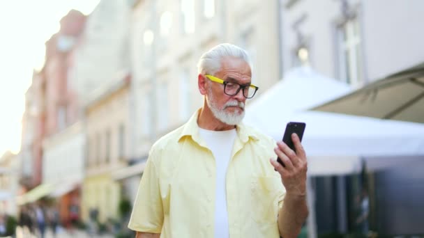 年长的白发苍苍的男人走在城市街道上浏览手机在线地图应用程序环视四周享受城市美景成熟的男性游客带着智能手机走到户外 — 图库视频影像