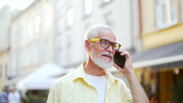 靠近点站在城市街道上的时候 一位头发灰白的大胡子老人正在用智能手机说话 微笑着穿着衬衫和眼镜的成熟的老男人 与同事或朋友交谈 — 图库视频影像