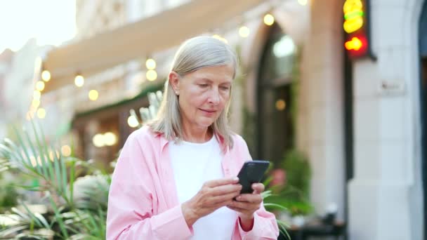 Mujer Pelo Gris Senior Utiliza Teléfono Inteligente Pie Una Calle — Vídeo de stock