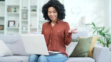 Genç Afro-Amerikalı iş kadını ev ofisindeki koltukta dizüstü bilgisayarla konuşuyor. Siyahi kadın serbest çalışan internetteki toplantıda iletişim kuruyor, webinar, müşteriyle istişare yapıyor.