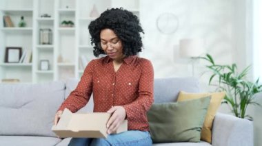 Heyecanlı bir Afrikalı Amerikalı kadın müşteri bir paket ev açıyor. Kanepede oturan mutlu siyah kadın alıcı paketleri açıyor. E-ticaretin tatmin olmuş müşterisi. Ekspres dağıtım hizmetleri kavramı