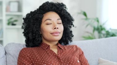 Genç, Afro-Amerikan bir kadının oturma odasında otururken gözleri kapalı dinlenmesine yakından bakın. Sakin ve mutlu bir siyahi kadının portresi evde derin derin nefes alıyor. Meditasyon kavramı