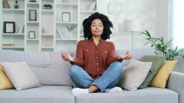 Genç Afro-Amerikan bayan meditasyon yapıyor gözleri kapalı evde oturma odasındaki kanepede nilüfer pozisyonunda oturuyor. Sakin, mutlu siyah bir kadın yoga yapar, derin nefes alır, rahat bir kanepede dinlenir.